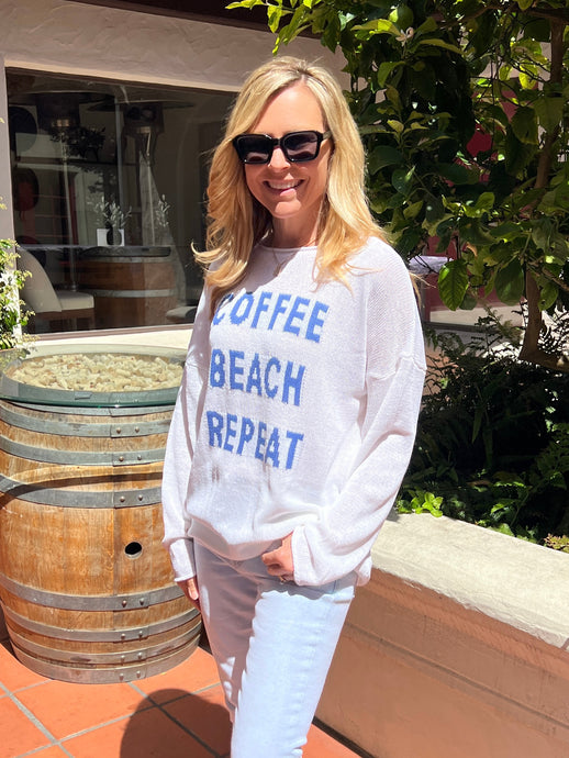 White sweater featuring the words “Coffee, Beach, Repeat” in bold blue on the front of the relaxed fit sweater.