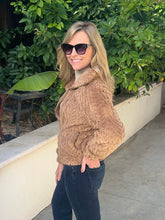 A woman wearing a brown cable-knit fuzzy cropped sweater, dark sunglasses, and dark jeans, standing outdoors in front of a beige wall and green foliage.