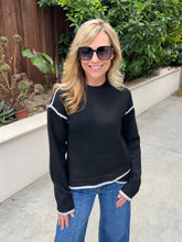 A woman wearing a black knit sweater with white contrast trim and blue jeans, standing outdoors on a concrete walkway with green plants and a wooden fence in the background.