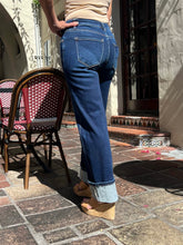 A person standing outdoors on a stone patio wearing dark blue cuffed jeans, a beige top, and wedge sandals. A wicker chair with a red-and-white checkered cushion is visible to the left.