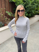 A woman wearing a light gray ribbed mock neck sweater, dark gray pants, and black sunglasses, standing outdoors on a concrete walkway with green plants and a wooden fence in the background.