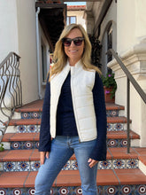 A woman wearing a white quilted sleeveless vest over a dark navy long-sleeve top and blue jeans, standing on tiled outdoor stairs.