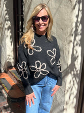 A woman wearing a dark charcoal knit sweater with large white floral patterns and light blue jeans, standing outdoors against a textured stucco wall with dappled sunlight and shadows.