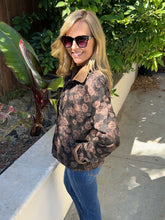A woman wearing a brown and beige floral print barn jacket with blue jeans, and sunglasses, standing outdoors on a concrete walkway next to a garden.