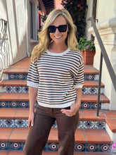 A woman wearing a brown and white striped short-sleeve knit sweater and brown pants stands on tiled outdoor steps in front of a building.