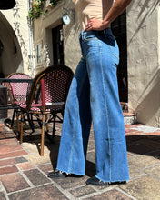 A woman wearing high-waisted, wide-leg blue denim jeans with a raw hem, styled with a beige tank top and black shoes, standing on a stone patio in front of a stucco building.