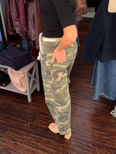 A person wearing camouflage cargo pants with a white drawstring waist, standing in a clothing store on a dark wood floor.