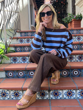 A woman wearing a blue and brown striped off-the-shoulder sweater, brown wide-leg pants, and tan wedge sandals sits on terracotta tiled steps with decorative blue and white patterned tiles.