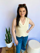 Woman wearing a cream sleeveless top and blue jeans standing next to a potted plant against a white wall.