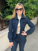 A woman wearing a dark blue denim jacket over a cream turtleneck sweater and dark jeans, standing outdoors in front of green foliage and a beige wall.