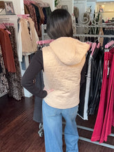 A woman viewed from behind wearing a quilted light beige hooded vest over a black long-sleeve top and blue jeans, standing inside a clothing store in front of racks of garments.