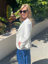 Woman wearing a white blazer and blue patterned dress standing outdoors with greenery in the background.