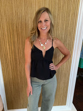 Woman wearing a black top and light olive jeans standing against a wooden paneled wall.