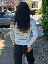 A woman viewed from behind wearing a fitted white long-sleeve top with thin gray horizontal stripes and black pants, standing outdoors on a sidewalk near parked cars and a building.