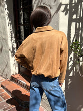 A person viewed from behind wearing a tan suede or nubuck button-up shirt and blue jeans, standing outdoors against a textured stucco wall with shadows.