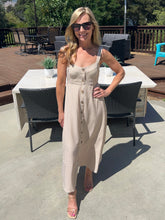Woman in a beige dress standing outdoors on a patio with furniture and plants.