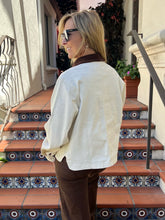 A woman wearing a cream-colored linen shirt and brown pants stands on tiled outdoor stairs, viewed from behind.