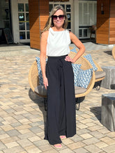 Woman in a white sleeveless top and black wide-leg pants standing outdoors on a patio.