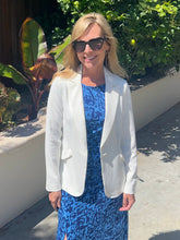 Woman in a white blazer and blue dress standing outdoors with greenery in the background