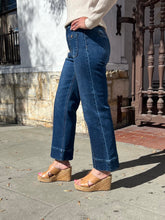 A person wearing dark blue straight-leg jeans with rolled cuffs, styled with tan wedge sandals and a cream ribbed sweater, standing outdoors on a sidewalk.