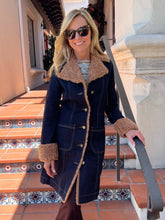 A woman wearing a dark navy blue denim-style coat with a tan shearling-style collar, cuffs, and front placket, standing on outdoor tiled steps.