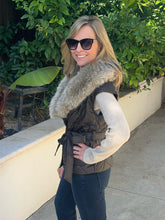 A woman wearing a dark brown quilted vest with a large beige faux-fur collar, a cream ribbed knit sweater, and dark blue jeans, standing outdoors on a concrete walkway with green foliage in the background.