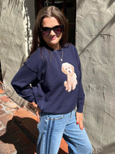A woman wearing a navy blue crewneck sweatshirt with a large beige and white poodle graphic on the front, paired with light blue jeans, standing outdoors against a textured gray wall.