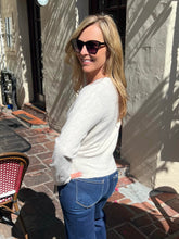 A woman wearing a light gray knit sweater and blue jeans stands outdoors on a sunlit patio, looking back over her shoulder. She is wearing dark sunglasses, with a stucco wall and patio furniture in the background.