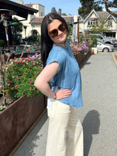 Woman wearing a chambray henley top and sunglasses standing outdoors with plants and buildings in the background.