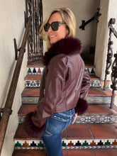 A woman wearing a brown faux-leather jacket with a dark burgundy faux-fur collar and matching fur pom-poms, paired with blue jeans and black sunglasses, posing on a tiled staircase.