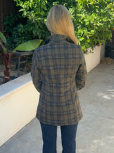 A woman viewed from behind wearing a brown and beige plaid flannel shirt jacket, standing on a concrete patio with green garden plants in the background.