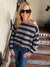 A woman wearing a gray and navy blue horizontally striped off-the-shoulder sweater, blue jeans, and black sunglasses, standing on an indoor tiled staircase.