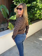 A woman wearing a brown long-sleeve knit sweater with black polka dots and blue jeans, standing outdoors on a concrete patio with green plants and a wooden fence in the background.