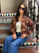 A woman wearing a brown and beige plaid cropped jacket over a cream tank top and blue jeans, sitting on a tiled bench with a decorative metal railing.