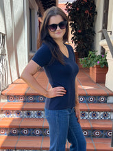 A woman wearing a navy blue short-sleeve V-neck t-shirt and blue jeans stands on tiled terracotta steps outdoors, wearing dark sunglasses.