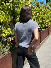A person standing outdoors in a garden setting, viewed from behind, wearing a textured short-sleeve gray top and black pants.