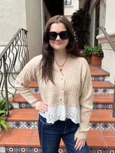 A woman wearing a beige cable-knit cardigan with a white eyelet lace hem, paired with dark blue jeans and black sunglasses, standing on terracotta tiled stairs.
