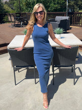Woman in a blue dress standing outdoors on a patio with tables and chairs.