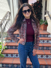 A woman posing on tiled outdoor stairs wearing a red and black tweed jacket over a burgundy top and blue jeans.