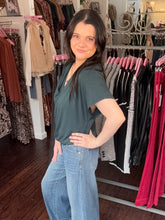 A woman with long dark hair stands in a clothing boutique wearing a dark green short-sleeve V-neck top and light blue wide-leg jeans, with racks of clothes behind her.
