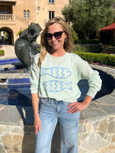 Woman wearing a light green sweater with fish designs and blue jeans standing in front of a fountain and building.
