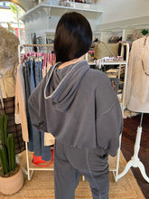 A person viewed from behind wearing a dark gray cropped hoodie with a silver zipper detail along the hood and matching dark gray sweatpants, standing inside a clothing boutique.