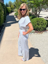Woman in a light outfit standing on a sidewalk with trees and shrubs in the background