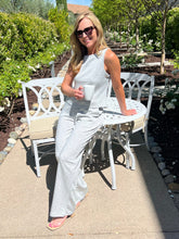 Woman in a white outfit sitting outdoors holding a cup, surrounded by greenery.