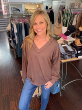A smiling blonde woman wearing a brown ribbed long-sleeve V-neck top and blue jeans, standing inside a clothing boutique with racks of garments and a table in the background.