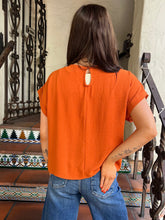 A woman viewed from behind wearing a bright orange short-sleeve top with a keyhole cutout at the back, paired with blue jeans, standing on tiled steps.