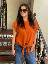 A woman wearing a bright orange short-sleeve knit top tied at the waist with blue jeans, standing on outdoor tiled steps.