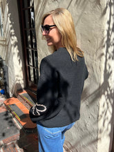 A woman with blonde hair and black sunglasses, wearing a dark charcoal gray knit sweater with a white beaded bow detail on the sleeve, paired with blue jeans, standing outdoors against a textured stucco wall.