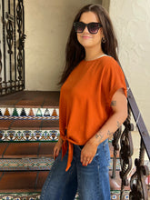 A woman wearing a rust-orange short-sleeve knit top with a side tie and blue jeans, standing on tiled stairs with a decorative wrought-iron railing.