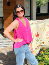 Woman in a pink top and blue jeans standing outdoors with a decorative chair and plants in the background.